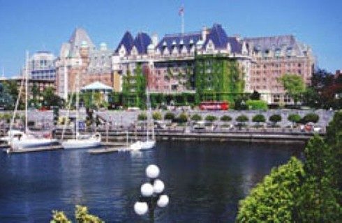 Large city buildings next to a harbor with tall white sailboats and row of lush green trees