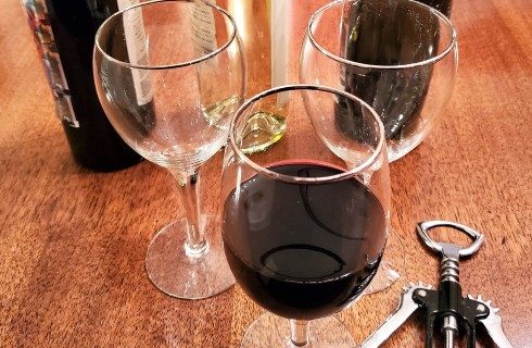 Three wine glasses; one filled with red wine, on a brown table with three bottles of wine and a bottle opener