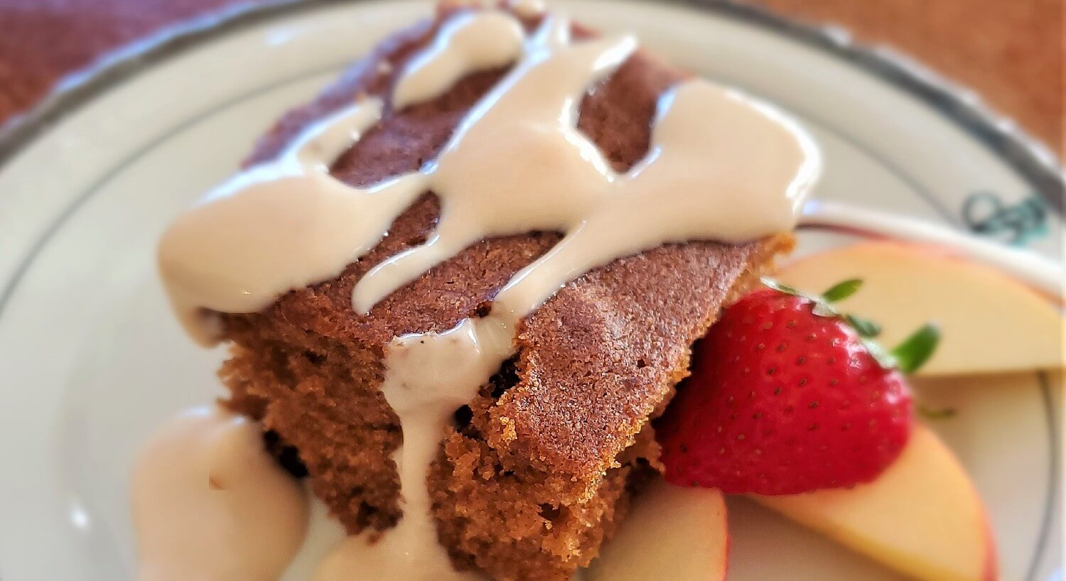 White plate with a square of coffee cake drizzled with white frosting and cut up strawberry and apples