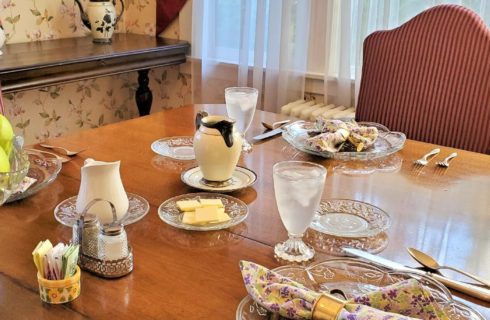 Dining table with upholstered chairs, set with glass plates holding flowered napkins in front of a window