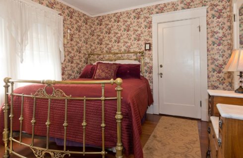 Queen bed with antique brass frame in bedroom with flowered wallpaper and large bright window