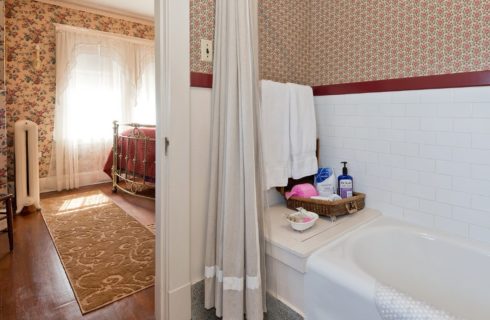 Bathroom with tub and tiled wall with view into bedroom with queen bed, wallpapered walls and bright windows