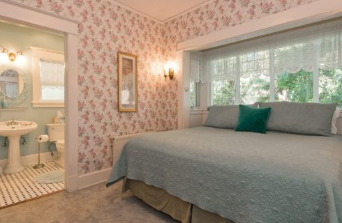 Large bedroom with flower wallpaper, king bed in front of windows and view into bathroom with pedestal sink