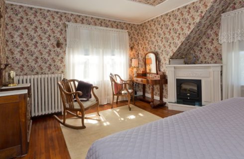 Corner sitting area of a bedroom with two wooden chairs, fireplace, vanity with mirror and large windows