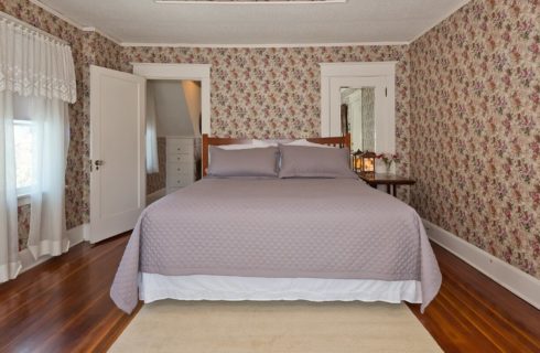 Large guest room with hardwood floors, flowered wallpaper, and king bed with lilac quilt