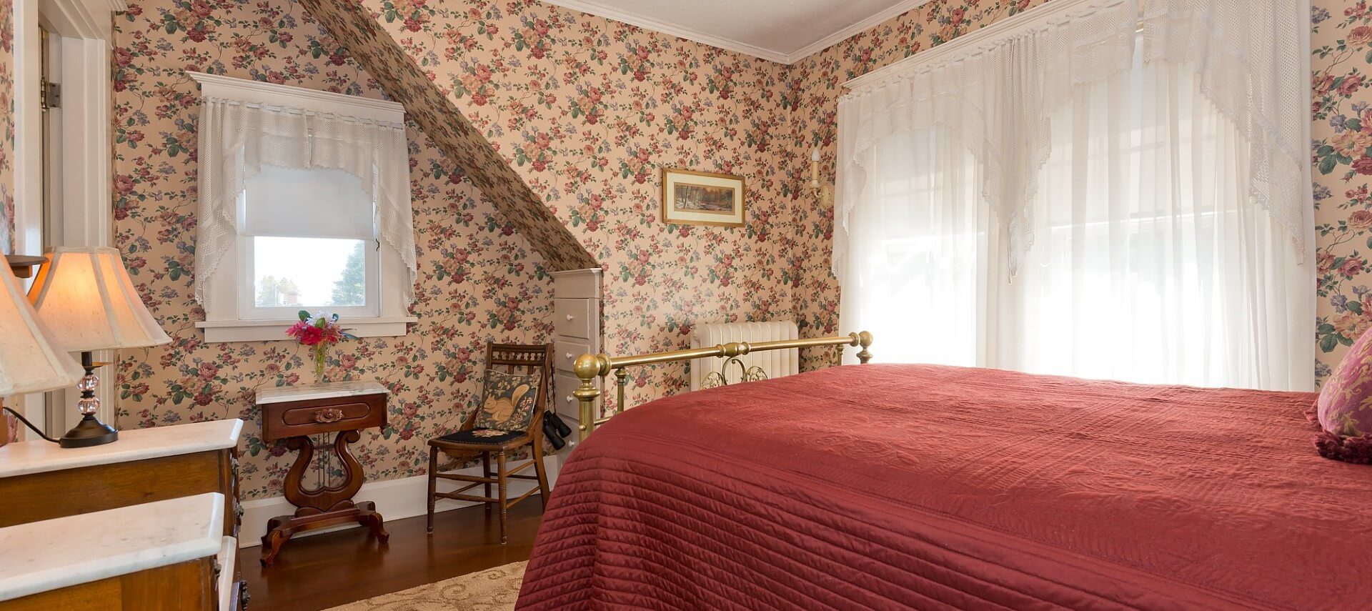 Queen bed with red quilt and brass frame in bedroom with flowered wallpaper and bright windows