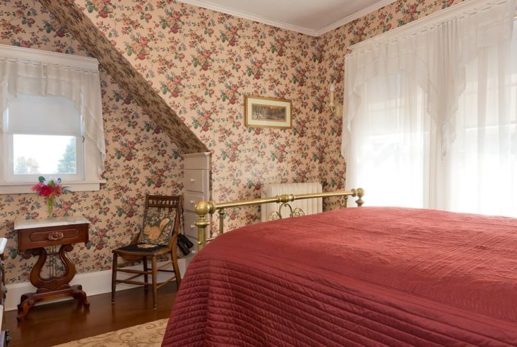 Queen bed with red quilt and brass frame in bedroom with flowered wallpaper and bright windows