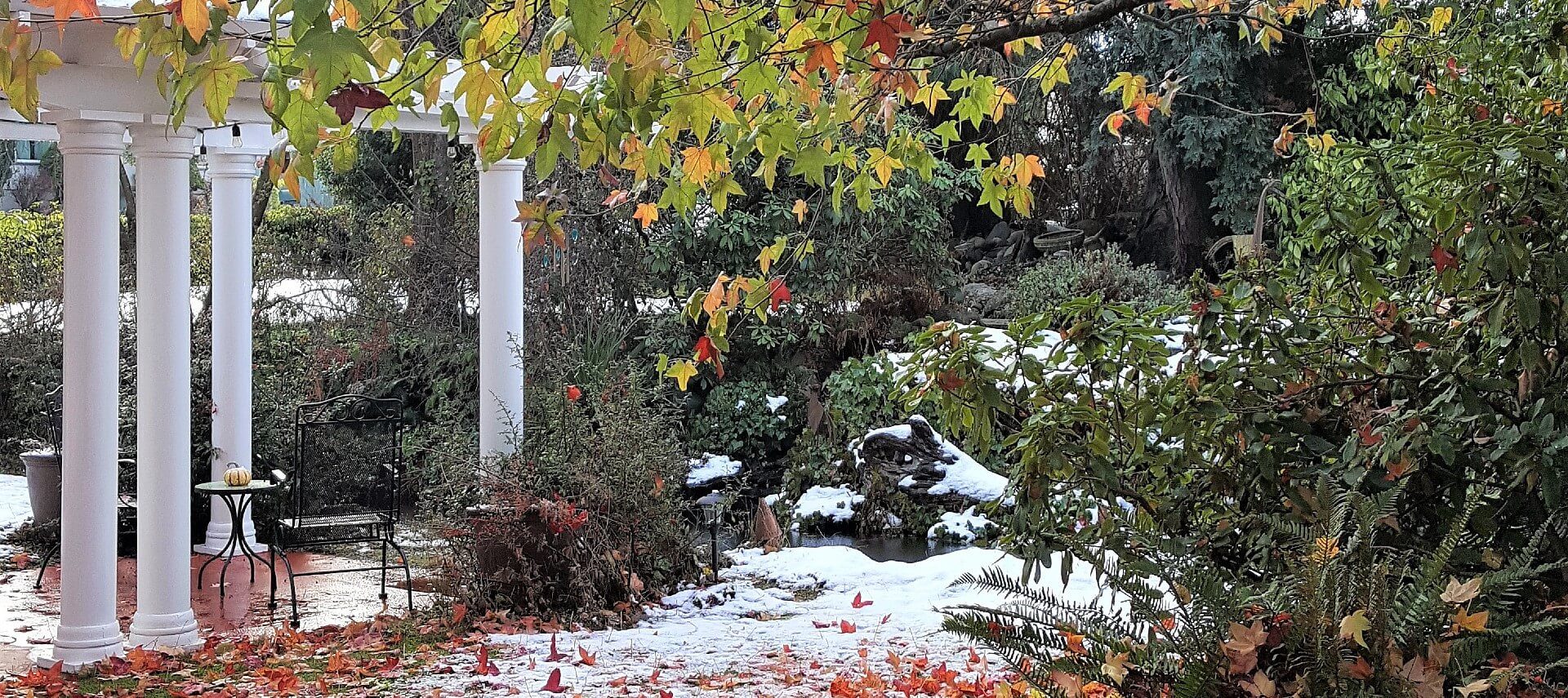 Outside garden with fall leaves and a bit of snow with patio and white pergola with columns