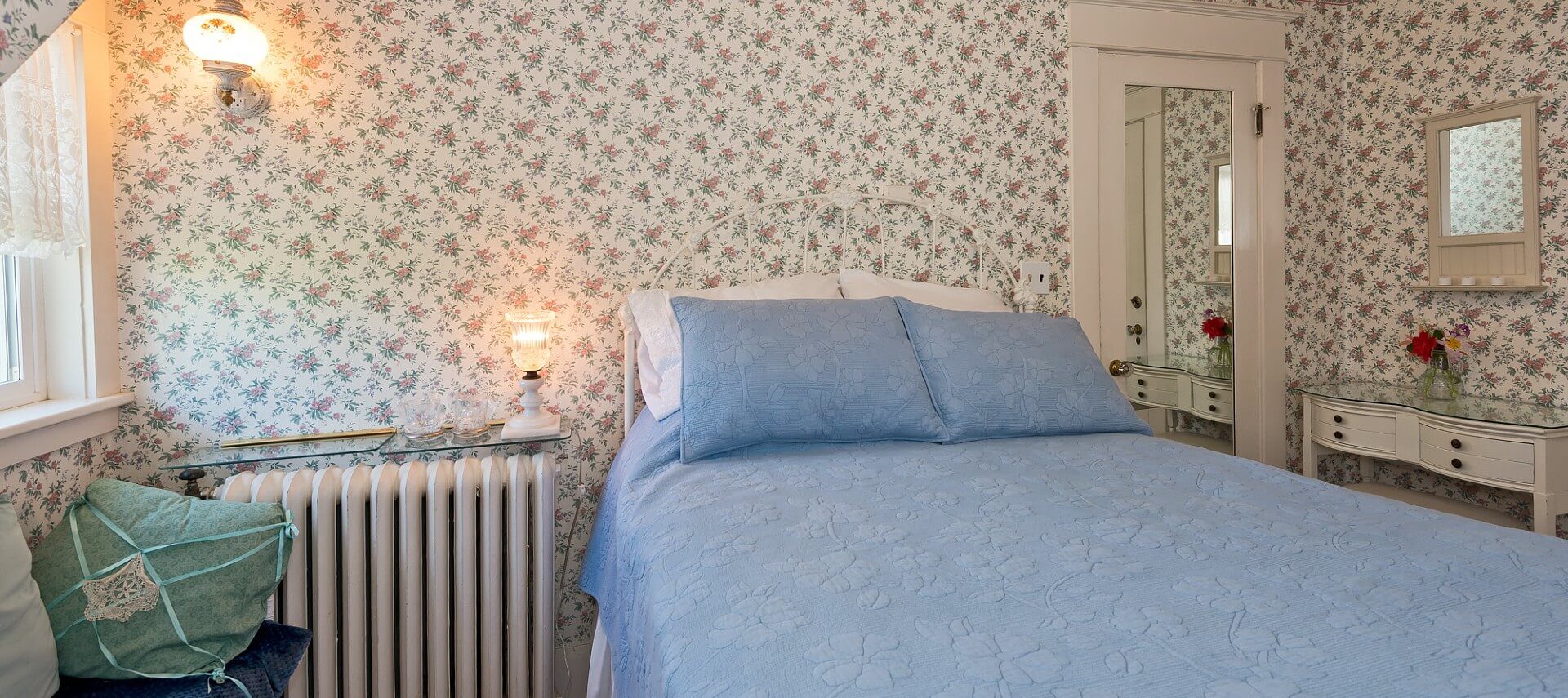 Guest room with flowered wallpaper, bright window and double bed with blue quilt