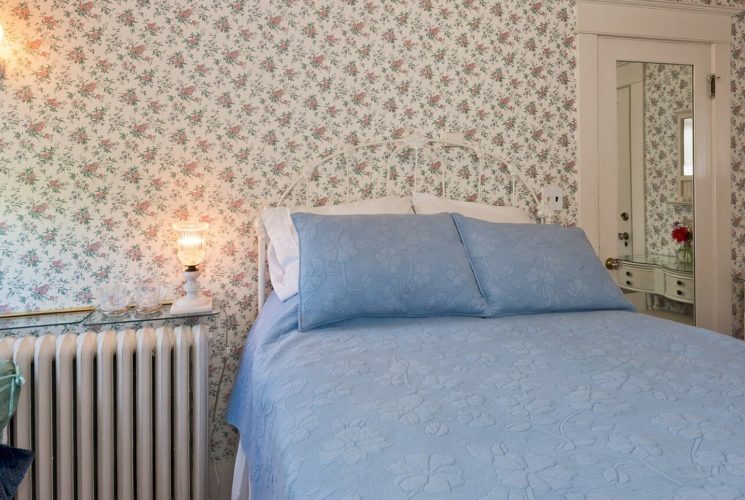 Guest room with flowered wallpaper, bright window and double bed with blue quilt