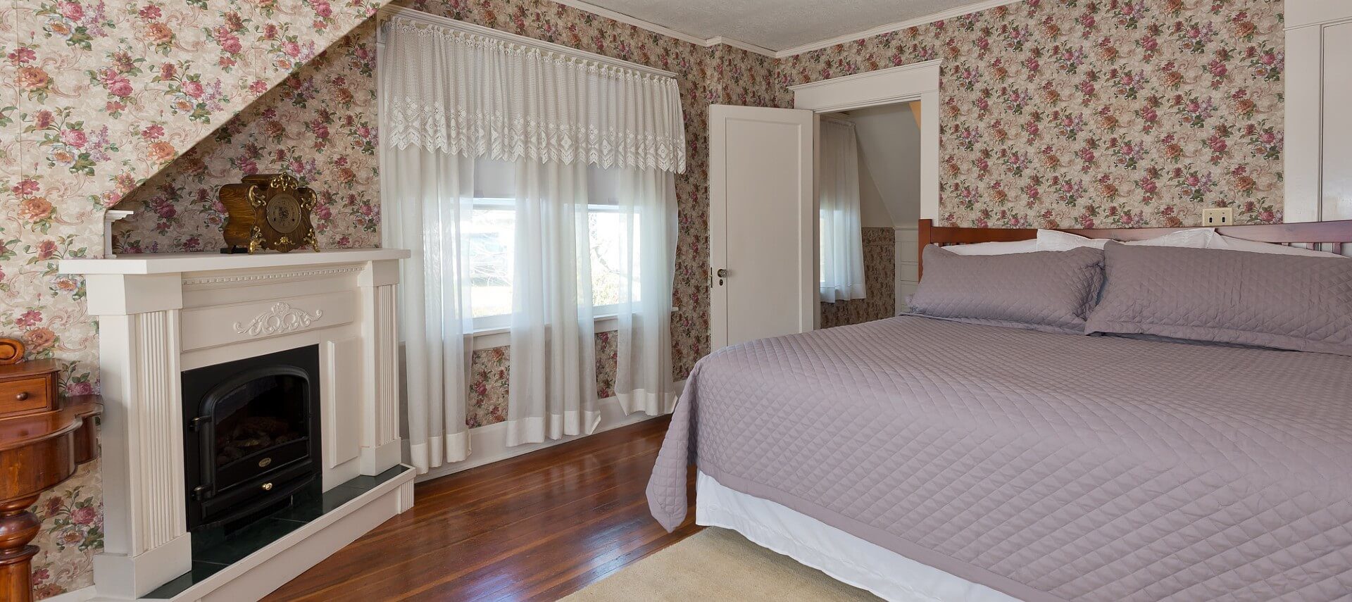 Large guest room with hardwood floors, wallpapered walls, fireplace and king bed with lilac quilt