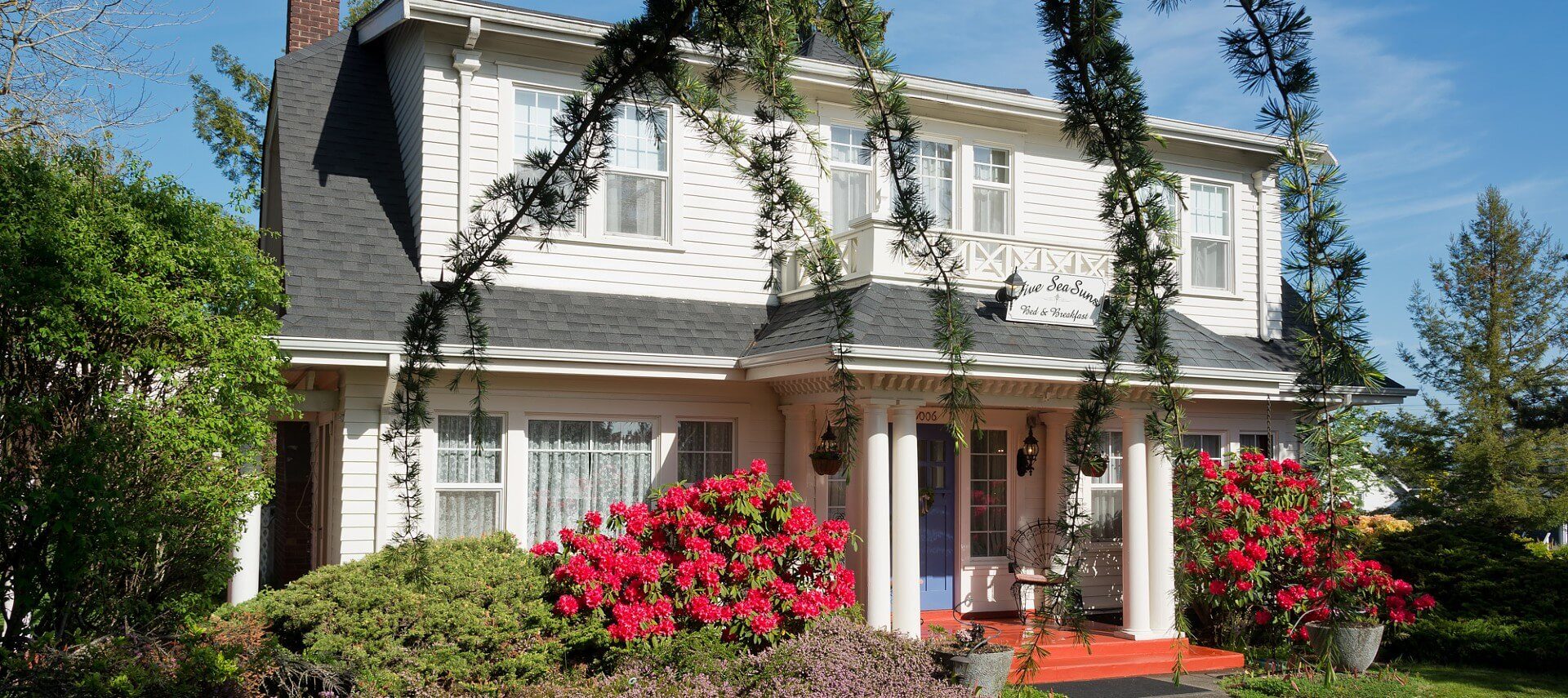 Grand white two story home with front porch, several windows and flowering bushes