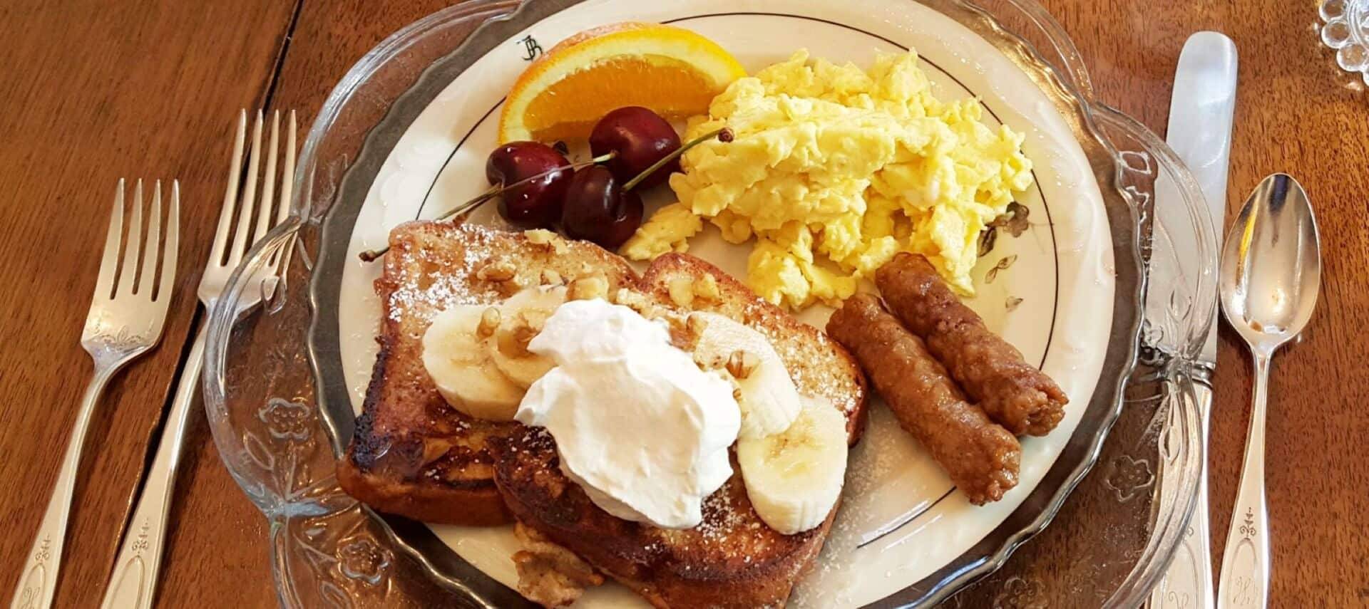 Glass and white plate showing french toast with bananas and whipped cream, sausage links, eggs and fruit