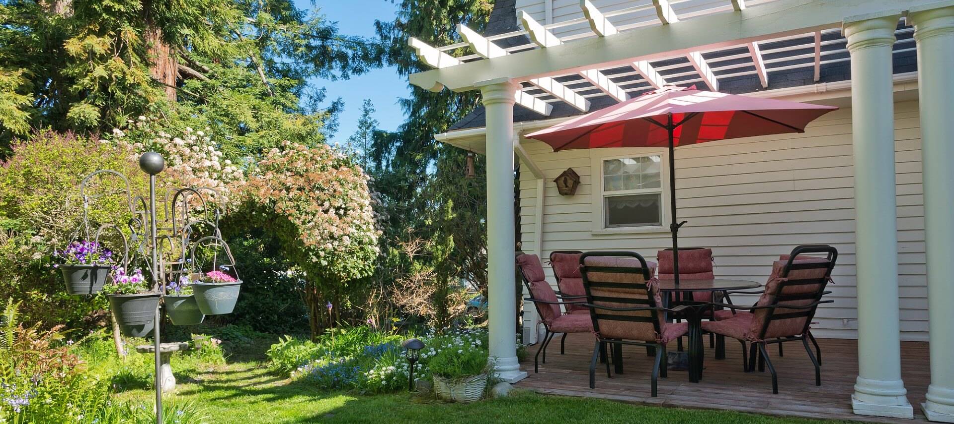 Outdoor patio with table, chairs and umbrella under a white pergola next to lush garden area